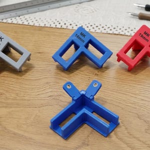 Puede incluir: Cuatro herramientas de sujeción de esquina en forma de L en gris, azul y rojo. La herramienta gris está marcada como MK 3/4", la azul como MK 16mm y la roja como MK 19mm. Una cuarta abrazadera de esquina azul tiene dos pequeños orificios circulares.