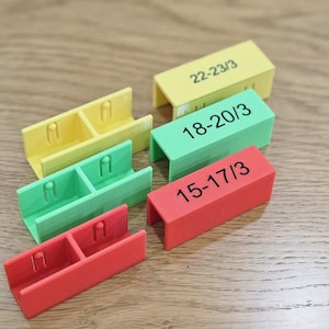 Puede incluir: Una colección de organizadores coloridos impresos en 3D en amarillo, verde y rojo. Cada organizador tiene forma rectangular con una ranura y los números "22-23/3", "18-20/3" y "15-17/3" impresos.
