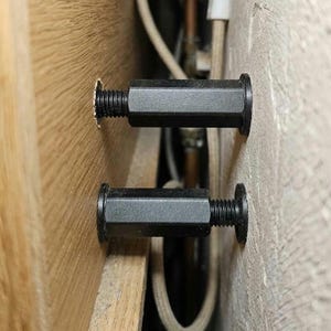Könnte beinhalten: Zwei schwarze Gewindebolzen sind zwischen einer Holzoberfläche und einer Betonwand installiert. Die Bolzen haben einen Sechskantkopf und einen kreisförmigen Flansch. Die Bolzen dienen zum Sichern oder Befestigen.