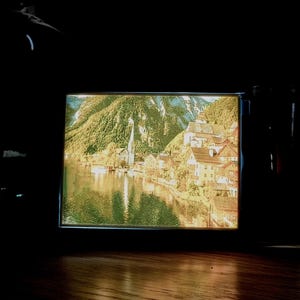 May include: A framed landscape print depicting a lakeside village nestled at the base of lush green mountains. The print features a church steeple and buildings with brown roofs, reflecting in the water.