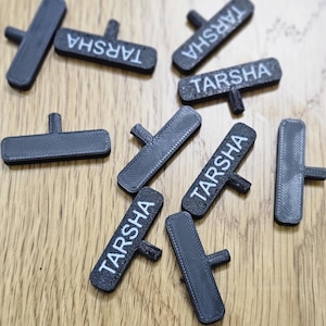 May include: A collection of black, rectangular name tags with a T-shaped handle. The word "TARSHA" is printed in white on each tag. The tags are scattered on a light brown wooden surface.