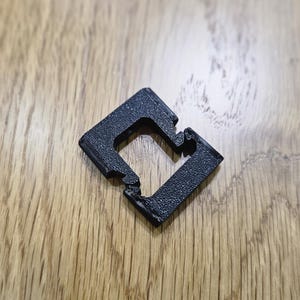 May include: A black, square-shaped 3D-printed object with a textured surface. The object has a central square opening and is resting on a wooden surface. The object appears to be a connector or a small component.