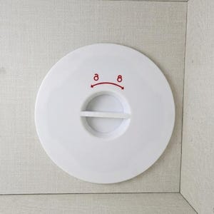 May include: A circular white plastic cover featuring a central horizontal bar handle. Above the handle, a red graphic shows two small circles connected by a curved arrow pointing in both directions, indicating a rotational locking mechanism.