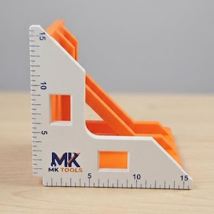 Op de afbeelding: Een L-vormig wit en oranje hoekklemgereedschap met een liniaalverdeling van 0 tot 15 cm op beide randen. Het gereedschap toont de tekst MK TOOLS met een moersleutelpictogram. Het bevat twee vierkante uitsparingen en een diagonale steunbalk.