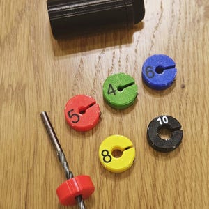Puede incluir: Una funda cilíndrica negra y un juego de topes para brocas de colores. Los topes son rojos, verdes, azules, amarillos y negros, cada uno con un número del 4 al 10. Se muestra una broca con un tope rojo adjunto.