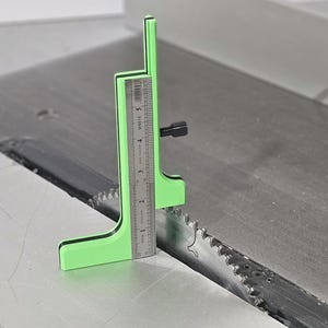 May include: A bright green woodworking tool with a ruler, measuring up to 5 inches. The tool is positioned over a saw blade, with a black adjustment knob. The saw blade is silver and has sharp teeth.