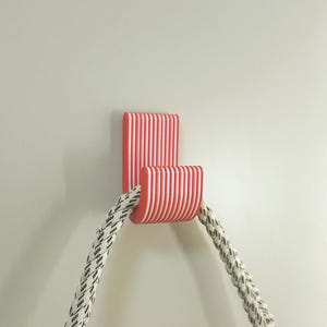 Pode incluir: Um gancho de parede listrado em vermelho e branco com um design curvo, segurando uma corda grossa com um padrão em preto e branco. O gancho é montado em uma parede lisa de cor clara.
