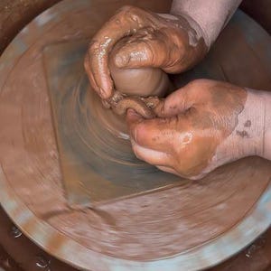 May include: A person's hands shaping a lump of clay on a pottery wheel. The clay is a light brown color and the wheel is spinning.