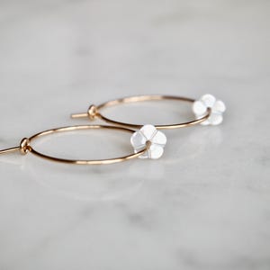 Puede incluir: Aretes de aro de oro con detalles de flores blancas. Los aretes son simples y elegantes.