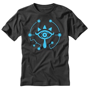 Puede incluir: Una camiseta negra con un gráfico azul y blanco del Ojo Sheikah del videojuego The Legend of Zelda: Breath of the Wild.