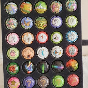 Puede incluir: Una colección de coloridos tapones de champán expuestos en una bandeja negra. Los tapones presentan varios diseños, incluyendo paisajes, arte abstracto y texto. Los tapones están dispuestos en un patrón de cuadrícula, mostrando una variedad de colores y estilos.