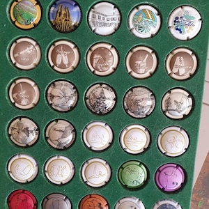 Puede incluir: Una colección de tapones de botellas de champán, cada uno con un diseño único. Los tapones están dispuestos en una vitrina verde. Los diseños incluyen paisajes, monumentos arquitectónicos e ilustraciones abstractas. Los tapones son de varios colores y estilos.