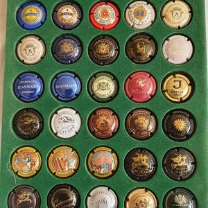 Puede incluir: Colección de tapones de botellas de champán expuestos en una bandeja verde. Los tapones presentan varios diseños, colores y nombres de marcas, incluyendo "Champagne Jeanmaire" y "Jacquart". Los tapones son redondos y metálicos, con logotipos y texto.