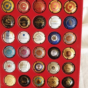 Puede incluir: Una colección de tapones de botellas de champán en varios colores, incluyendo dorado, rojo y plateado, exhibidos en una bandeja roja. Los tapones presentan nombres de marcas y diseños decorativos, mostrando una variedad de marcas de champán.
