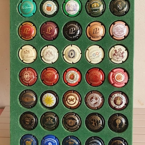 Puede incluir: Una colección de 49 tapones de botellas de champán expuestos en un marco forrado de fieltro verde. Los tapones están dispuestos en filas y columnas, y cada tapón presenta un diseño diferente, incluidos logotipos, escudos de armas y texto. Los tapones son una mezcla de oro, plata y rojo.