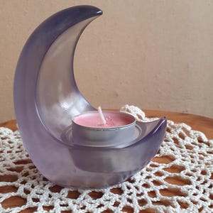 Peut inclure: Un bougeoir en forme de croissant de lune violet translucide fait d'un matériau lisse. Le bougeoir présente une découpe circulaire au centre conçue pour maintenir solidement une petite bougie chauffe-plat ronde à cire rose et mèche blanche.