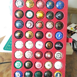 Puede incluir: Una caja de terciopelo rojo con 49 tapones de botellas de champán. Los tapones están dispuestos en una cuadrícula y presentan varios logotipos y diseños. Algunos de los logotipos incluyen "Ruinart", "Lanson", "Moët & Chandon" y "Champagne M.C."