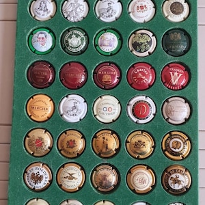 Puede incluir: Colección de tapones de botellas de champán en un expositor verde. Los tapones redondos presentan diversos diseños, logotipos y textos en dorado, rojo, negro y blanco. Dispuestos en filas y columnas.