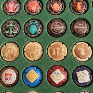 Puede incluir: Colección de tapones de botellas de champán con varios diseños y colores. Algunos tapones presentan símbolos del zodiaco, mientras que otros muestran ilustraciones de personas y paisajes. Los tapones están dispuestos en una vitrina verde.