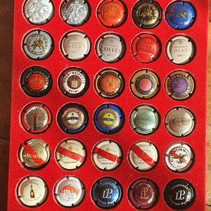 Puede incluir: Una colección de 49 tapones de botellas de champán expuestos en una caja forrada de terciopelo rojo. Los tapones están dispuestos en filas y columnas, y cada tapón presenta un nombre de marca y un diseño diferentes. Algunas de las marcas incluyen Moët & Chandon, Veuve Clicquot y Laurent-Perrier.