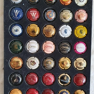 Puede incluir: Una colección de tapones de botellas de champán, cada uno con un diseño y nombre de marca únicos, dispuestos en una vitrina negra. Los tapones presentan varios colores, logotipos y texto, mostrando una variedad de marcas de champán.