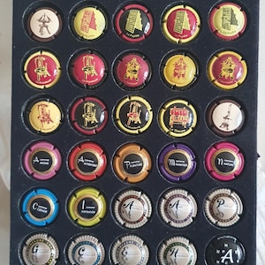 Puede incluir: Una colección de corchos de champán con varios diseños y textos, dispuestos en una caja expositora negra. Los corchos presentan ilustraciones coloridas y palabras como "Arome" e "Inspiration".
