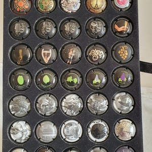 Puede incluir: Una colección de cápsulas de botellas de champán expuestas en una bandeja negra. Las cápsulas presentan varios diseños, incluyendo patrones geométricos, ilustraciones y texto. Algunas cápsulas tienen detalles dorados, plateados y coloridos.