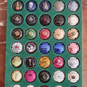 Puede incluir: Colección de tapones de botellas de champán expuestos en una bandeja forrada de fieltro verde. Los tapones presentan varios diseños, incluyendo ilustraciones y logotipos, con colores como dorado, rojo y negro. La bandeja está sobre una superficie de madera.