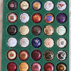 Puede incluir: Colección de tapones de botellas de champán en una vitrina verde. Los tapones son redondos, con varios colores y diseños, incluyendo texto y logotipos. Están dispuestos en un patrón de cuadrícula.
