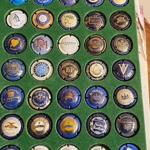 Puede incluir: Una colección de 36 tapones de botellas de champán expuestos en una bandeja verde cuadriculada. Los tapones son principalmente azules y dorados con varios logotipos y textos, incluyendo "Champagne Jeanmaire Epernay", "Champagne De Barfontarc", "Champagne Duval Leroy", "Champagne Besserat de Bellefon", "Champagne Bruno Paillard", "Champagne Vertus", "Champagne Veuve Clicquot", "Champagne Pommery", "Champagne Pol Roger & Co. Epernay", "Champagne Delamotte", y "Champagne AMF".