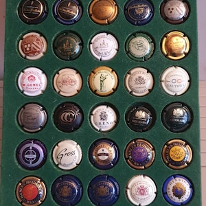Puede incluir: Una vitrina verde con 42 tapones de botellas de champán. Los tapones están dispuestos en forma de cuadrícula y presentan varios logotipos y diseños. Algunos de los logotipos incluyen "Nicolas Feuillatte", "M. Gonel Damery", "Gross", "Henriot" y "Herbert Heidsieck".