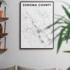 May include: A black and white map print of Sonoma County, California, in a black frame. The map shows the roads and towns of the county. The print is hanging on a white wall above a couch with pillows.