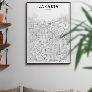 May include: A black and white map print of Jakarta, Indonesia, in a black frame. The map shows the city's streets and landmarks. The print is hanging on a white wall above a couch with pillows.