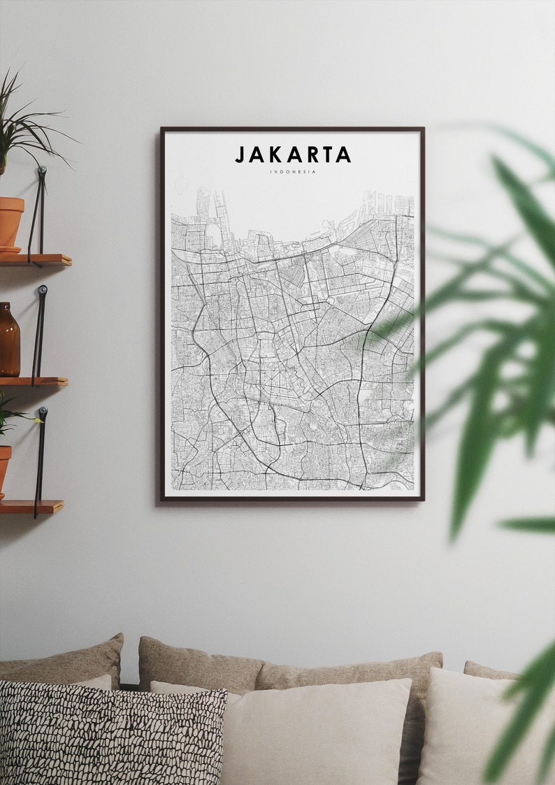 May include: A black and white map print of Jakarta, Indonesia, in a black frame. The map shows the city's streets and landmarks. The print is hanging on a white wall above a couch with pillows.