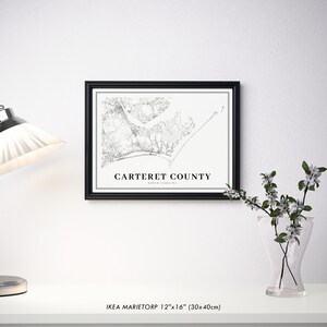 May include: A black framed print of a map of Carteret County, North Carolina. The map is in black and white and shows the roads, rivers, and coastline of the county. The text 'Carteret County, North Carolina' is printed below the map. The print is sitting on a white surface with a vase of white flowers and a lamp to the left.