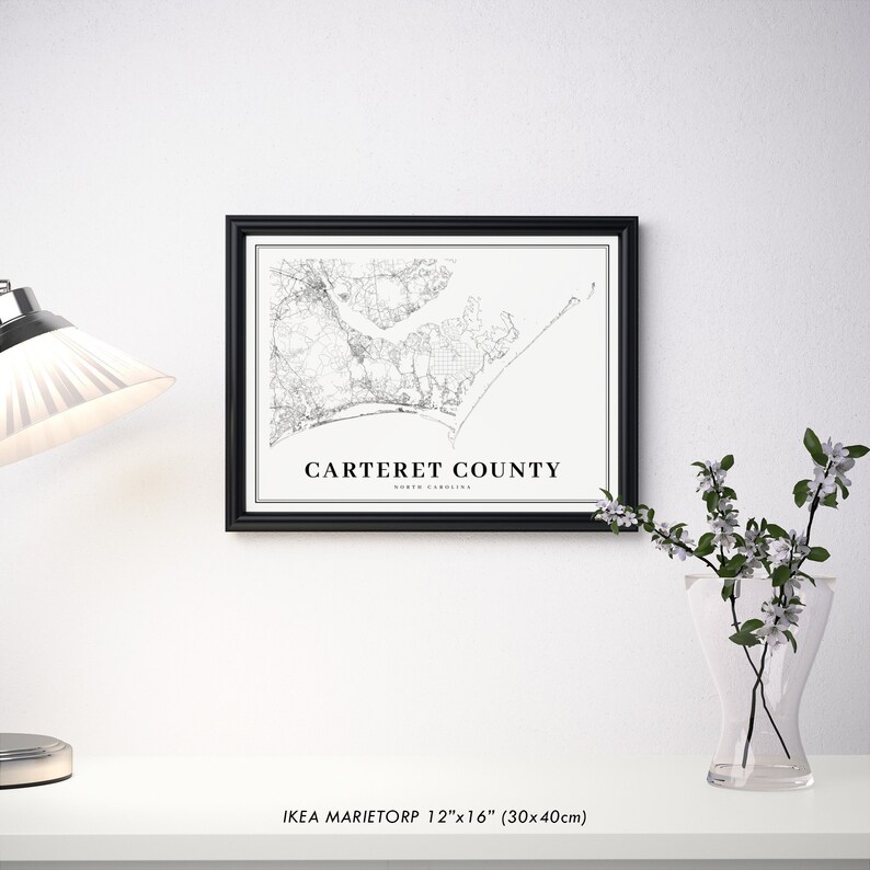 May include: A black framed print of a map of Carteret County, North Carolina. The map is in black and white and shows the roads, rivers, and coastline of the county. The text 'Carteret County, North Carolina' is printed below the map. The print is sitting on a white surface with a vase of white flowers and a lamp to the left.