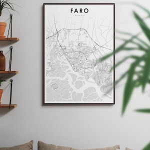 May include: A black and white map print of Faro, Portugal, in a black frame. The map shows the city's streets and waterways. The print is hanging on a white wall above a sofa with cushions.