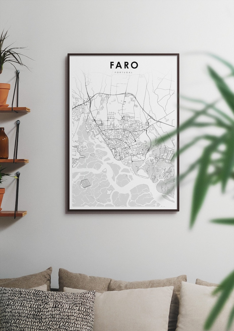 May include: A black and white map print of Faro, Portugal, in a black frame. The map shows the city's streets and waterways. The print is hanging on a white wall above a sofa with cushions.