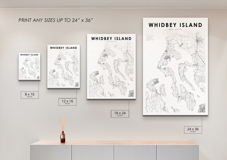 May include: Four different sizes of a black and white map print of Whidbey Island, Washington. The prints are displayed on a white wall with a light wood shelf underneath. The sizes are 8x10 inches, 12x16 inches, 18x24 inches, and 24x36 inches.