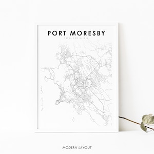 Op de afbeelding: Een zwart-witte kaartprint van Port Moresby, Papoea-Nieuw-Guinea, met de naam van de stad en de locatie gedrukt bovenaan de kaart. De kaart is ingelijst in een wit frame en wordt weergegeven op een witte muur.