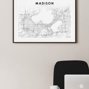 May include: A black and white map of Madison, Wisconsin, framed in a black frame, hanging on a white wall. A black office chair and a silver laptop are on a wooden desk in the foreground.