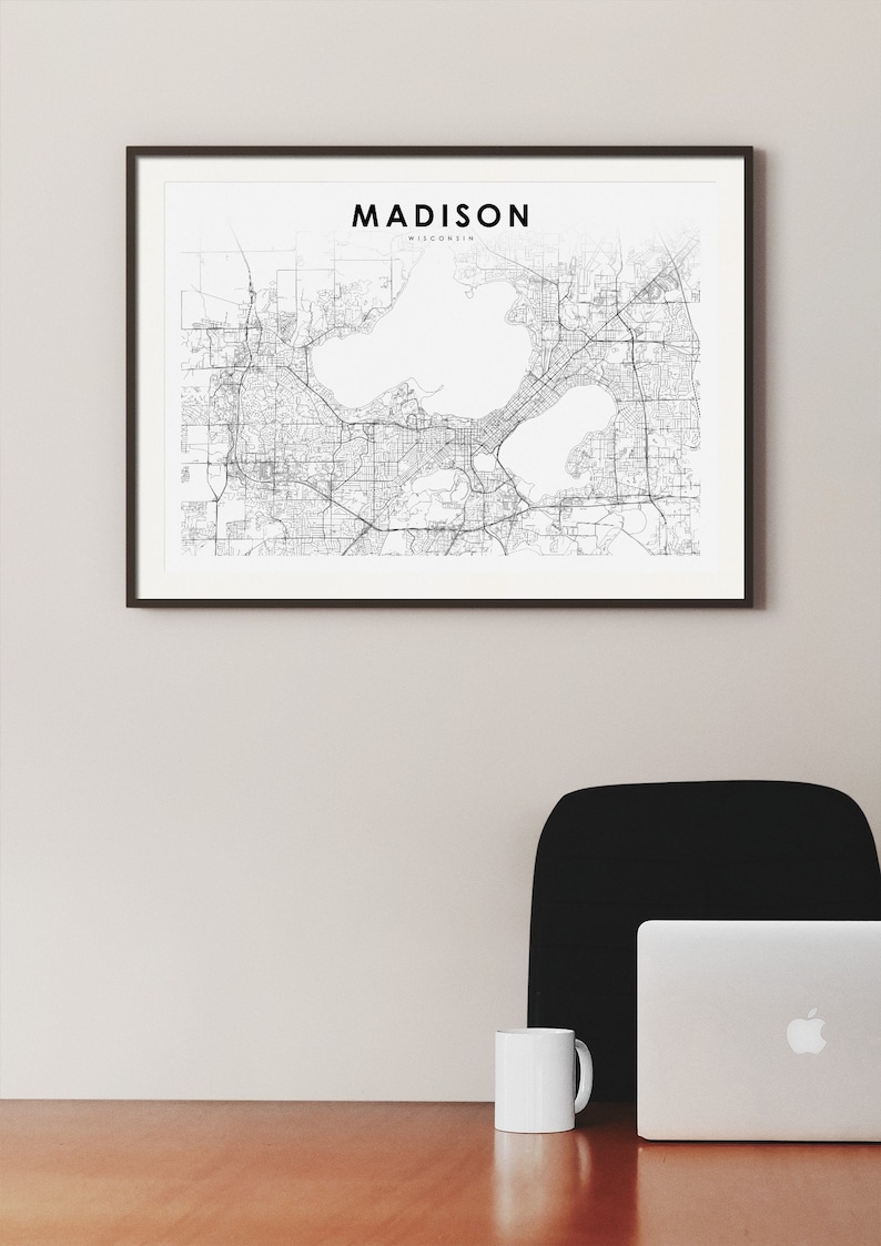May include: A black and white map of Madison, Wisconsin, framed in a black frame, hanging on a white wall. A black office chair and a silver laptop are on a wooden desk in the foreground.