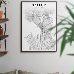 May include: A black and white map print of Seattle, Washington, in a black frame. The map shows the city's streets and waterways. The print is hanging on a white wall above a couch with pillows.