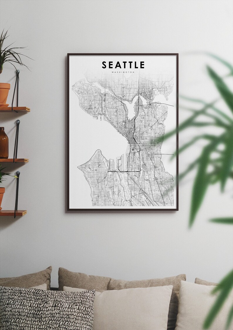 May include: A black and white map print of Seattle, Washington, in a black frame. The map shows the city's streets and waterways. The print is hanging on a white wall above a couch with pillows.