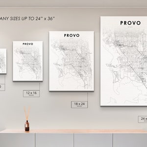 May include: A white wall with four black and white city maps of Provo, Utah, framed and hanging on the wall. The maps are in different sizes: 8x10 inches, 12x16 inches, 18x24 inches, and 24x36 inches. The text "PRINT ANY SIZES UP TO 24" x 36" is at the top of the wall. The maps are on a white wall above a light brown wooden shelf.