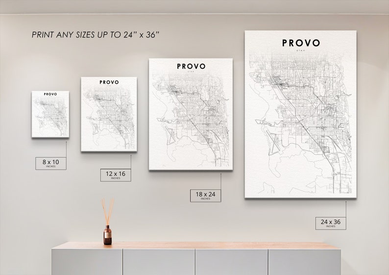 May include: A white wall with four black and white city maps of Provo, Utah, framed and hanging on the wall. The maps are in different sizes: 8x10 inches, 12x16 inches, 18x24 inches, and 24x36 inches. The text "PRINT ANY SIZES UP TO 24" x 36" is at the top of the wall. The maps are on a white wall above a light brown wooden shelf.