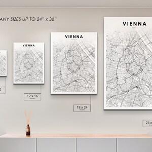 Op de afbeelding: Een witte muur met vier zwart-witte stadskaarten van Wenen, Oostenrijk, in witte lijsten. De kaarten zijn in verschillende formaten: 8 x 10 inch, 12 x 16 inch, 18 x 24 inch en 24 x 36 inch. De tekst "DRUK ALLE FORMATEN TOT 24" x 36" staat bovenaan de muur.