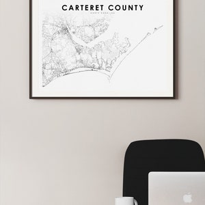 May include: A black and white map of Carteret County, North Carolina, framed in a black frame and hanging on a white wall. The map is surrounded by a white mat. The map is printed on white paper and shows the roads, rivers, and coastline of the county. The title of the map is 'Carteret County, North Carolina'.