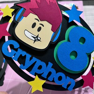 Pode incluir: Um topo de bolo preto e azul com um personagem de desenho animado usando fones de ouvido e o nome "Cryphon" e o n&uacute;mero "8". O topo &eacute; decorado com estrelas amarelas e vermelhas.