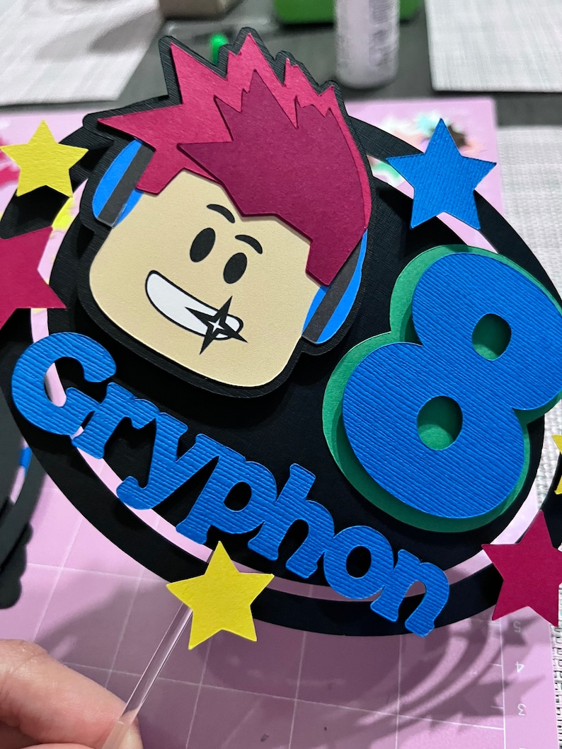 Pode incluir: Um topo de bolo preto e azul com um personagem de desenho animado usando fones de ouvido e o nome "Cryphon" e o n&uacute;mero "8". O topo &eacute; decorado com estrelas amarelas e vermelhas.